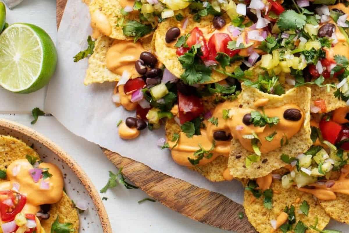 A plate of loaded nachos topped with melted cheese, black beans, diced tomatoes, chopped onions, cilantro, and other vegetables. Lime halves are placed nearby, adding color and suggesting a garnish. The dish is served on a round, wooden platter.