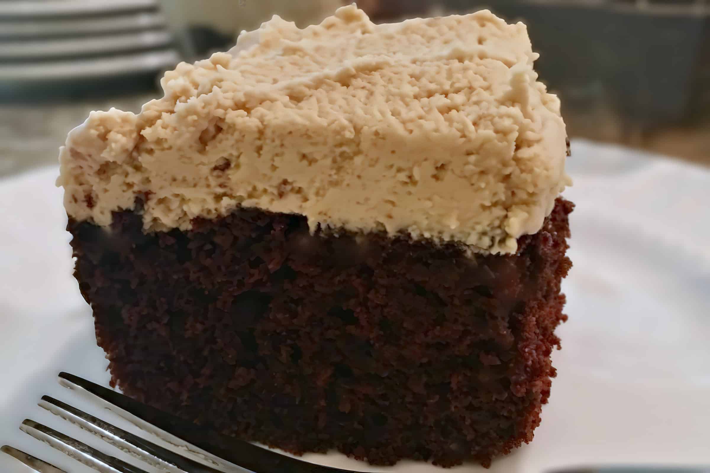 A close-up of a slice of chocolate cake topped with a thick layer of creamy frosting on a white plate. A fork is positioned to the left side of the plate, ready to be used. The background is blurred, keeping the focus on the cake.