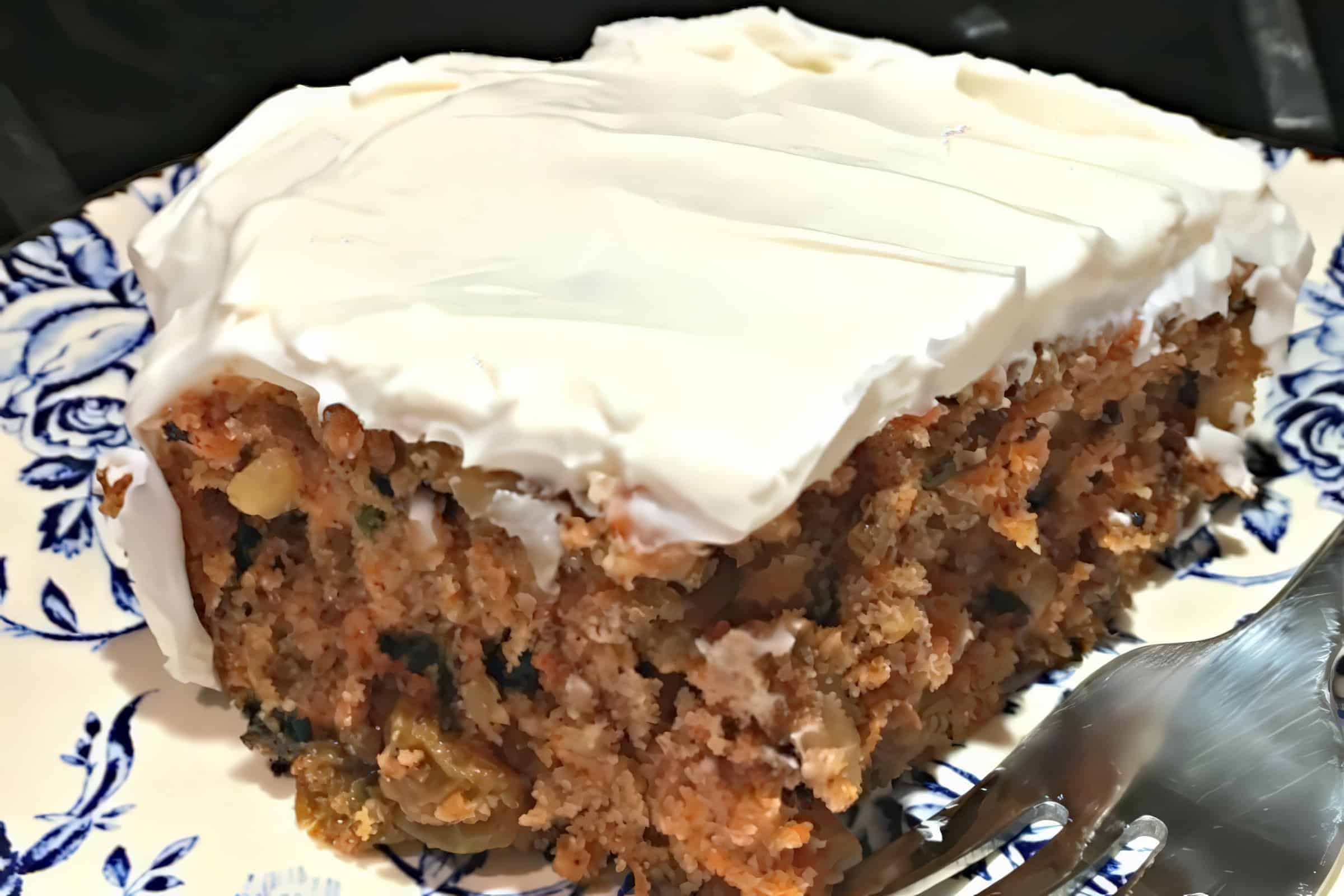 A slice of carrot cake sits on a plate with a blue floral pattern. The cake has a moist texture and is topped with a thick layer of creamy white icing. A metal fork to the lower right suggests it is ready to be enjoyed.