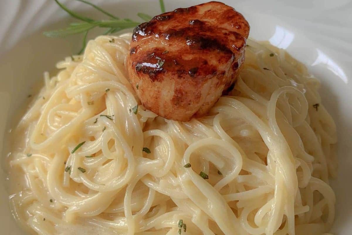 A close-up image of a creamy pasta dish topped with a seared scallop. The pasta is coated in a thick, creamy sauce and garnished with finely chopped herbs. The scallop is browned and glistening, placed at the center of the pasta mound.