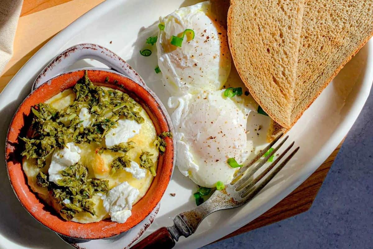 A white plate holding two slices of toasted bread, two poached eggs garnished with green onions, and a small dish of greens with crumbled cheese. A fork rests on the plate, and sunlight casts shadows on the meal.