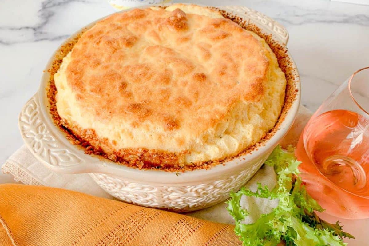 A baked dish with a golden-brown crust sits in an ornate round casserole. A glass of rosé and green salad leaves are placed next to an orange napkin on a white marble surface.