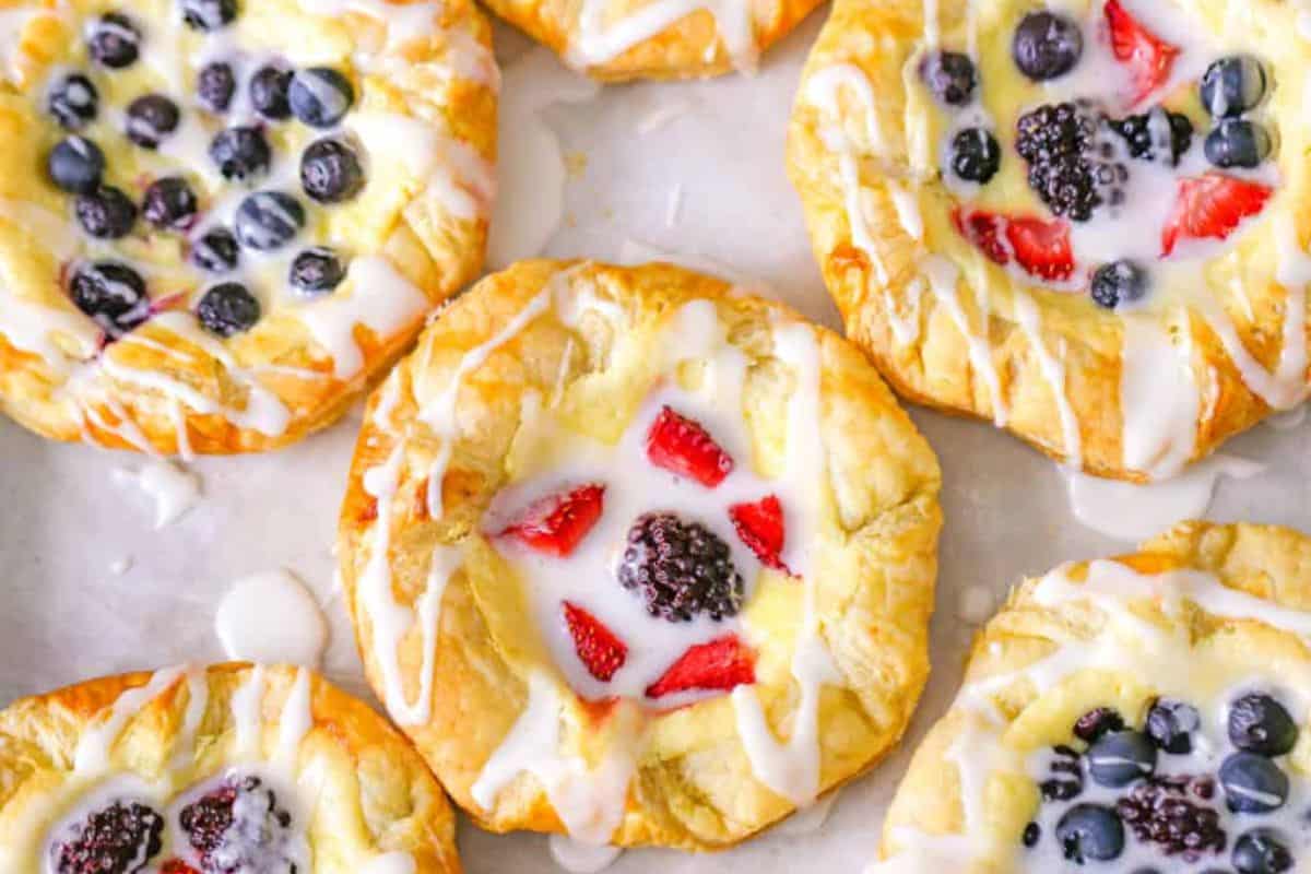 An assortment of fruit-topped pastries is displayed, each drizzled with a glaze. The pastries feature various fruits, including blueberries, strawberries, and blackberries, set on a cream-filled, golden-brown flaky dough. Several pastries are visible on a white surface.