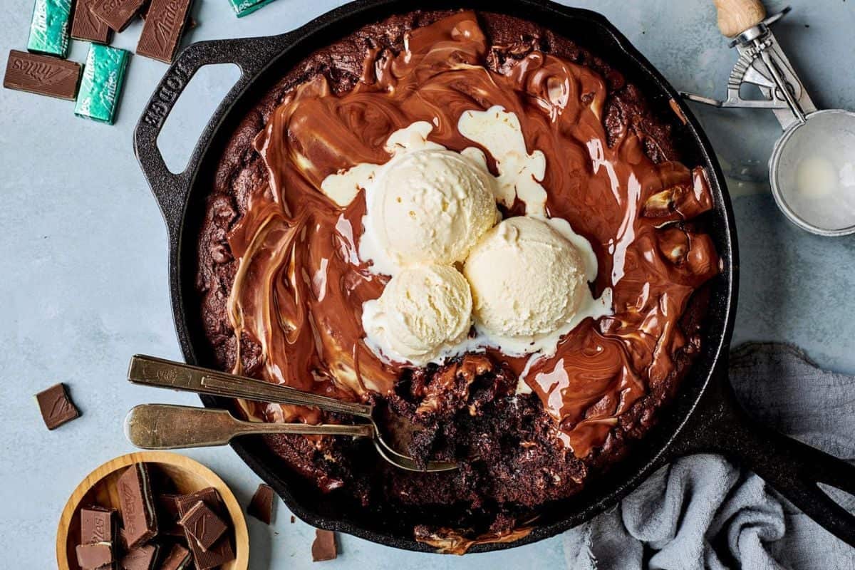 A cast iron skillet holds a gooey chocolate brownie topped with melted chocolate and three scoops of vanilla ice cream. Two spoons are placed in the brownie. Surrounding the skillet are pieces of chocolate, mint chocolate, and an ice cream scoop on a gray cloth.