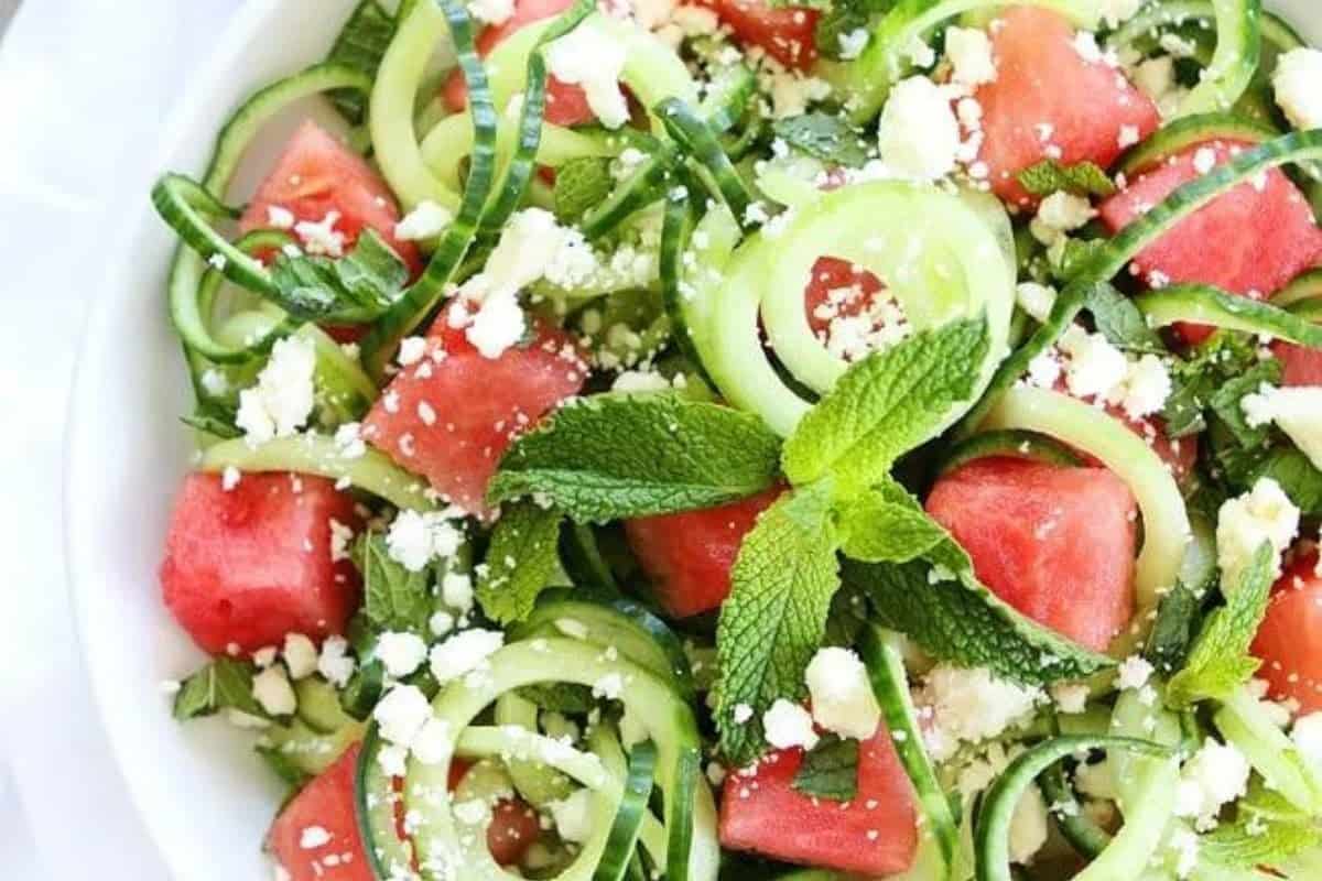 A fresh salad featuring coiled cucumber slices, chunks of watermelon, topped with crumbled feta cheese, and garnished with sprigs of mint leaves. The vibrant green, pink, and white colors highlight its refreshing and appetizing appearance.