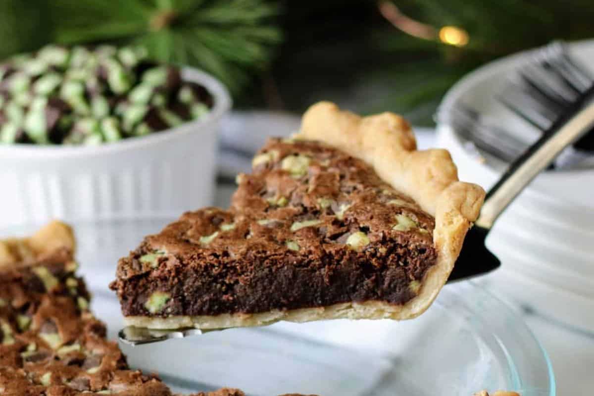 A slice of chocolate mint pie is being lifted from a pie dish. The pie has a thick, dark chocolate filling with green mint pieces and a golden crust. In the background, a bowl of mint-topped treats and a stack of white plates with forks are visible.