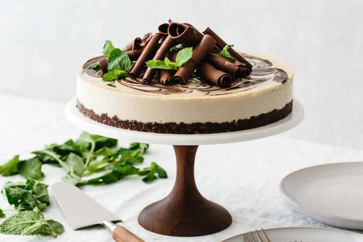 A cheesecake with a chocolate crumb crust on a wooden cake stand, topped with chocolate curls and fresh mint leaves. A cake knife and sprigs of mint are placed on the white tablecloth next to it, with a couple of empty plates nearby.