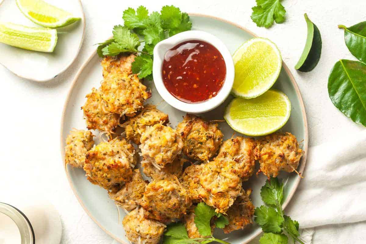 A plate of crispy crab cakes garnished with cilantro, accompanied by a small bowl of red dipping sauce and lime wedges on the side. The setup is placed on a white surface with some leaves scattered around.