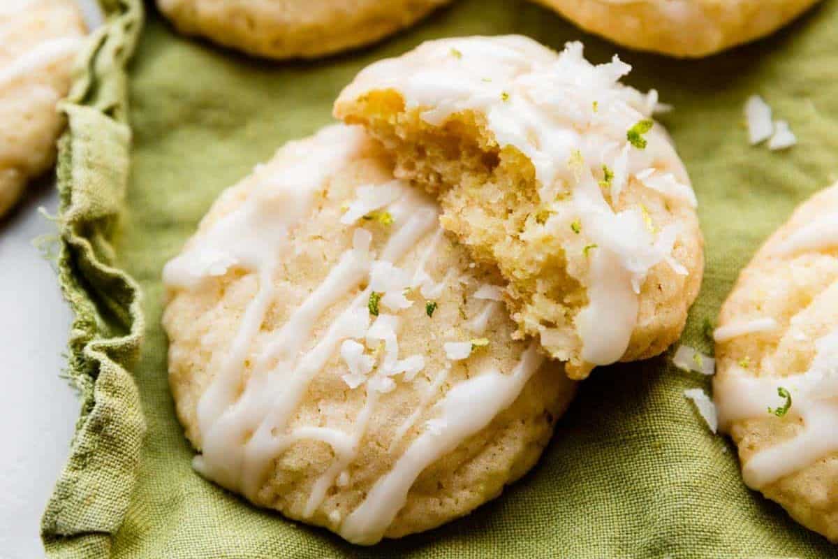 A close-up of a cookie with a bite taken out of it, revealing its soft interior. The cookie is drizzled with white icing and sprinkled with small bits of coconut and lime zest. It rests on a green cloth. Other cookies are partially visible in the background.