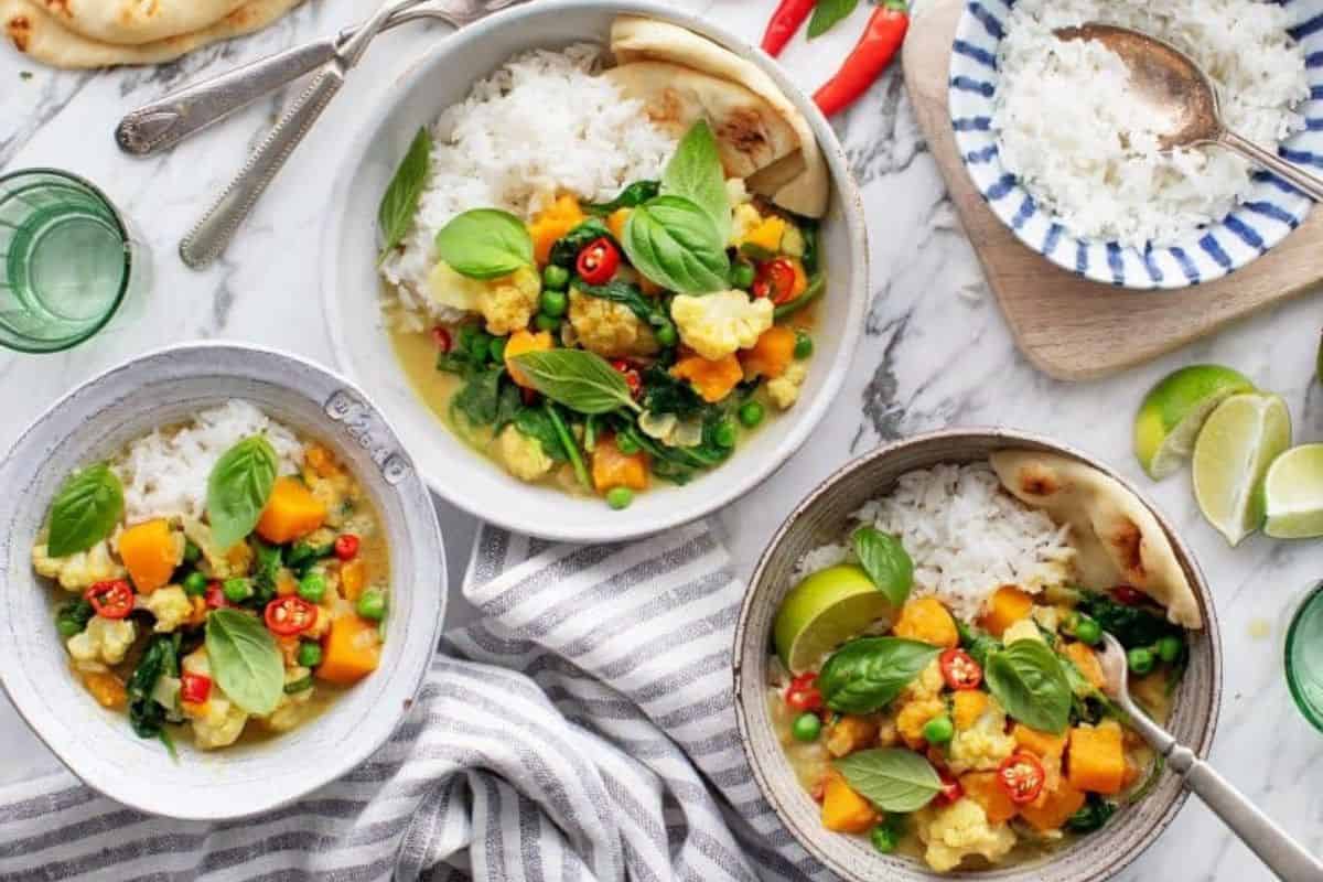 A colorful spread of vegetable curry dishes with white rice, garnished with fresh basil leaves and red chili slices. Two bowls and a plate are presented on a marble surface, accompanied by lime wedges, glassware, and pieces of bread.