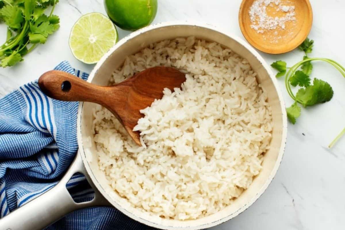 A pot of cooked white rice with a wooden spoon placed inside. Next to the pot are fresh lime halves, a wooden bowl of coarse salt, cilantro leaves, and a blue-striped cloth napkin on a marble surface.