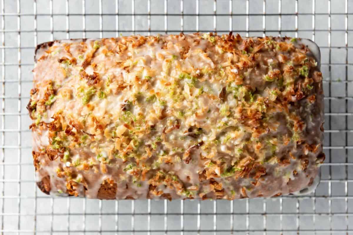 A freshly baked loaf of bread, covered in a light glaze and sprinkled with grated lime zest and coconut, sits on a cooling rack. The loaf has a golden-brown crust and is placed against a white background.