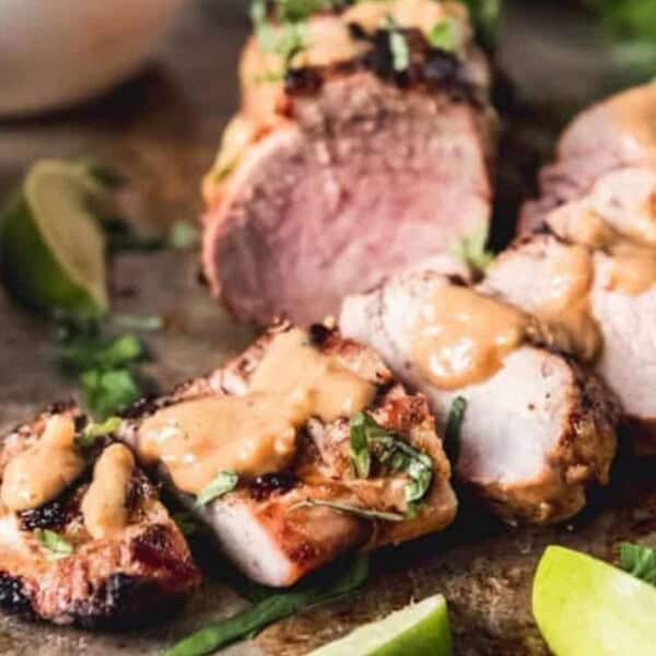 A close-up view of sliced grilled pork tenderloin topped with a creamy mustard sauce, garnished with fresh herbs. Lime wedges and a small bowl of extra sauce are placed beside the tenderloin on a rustic wooden surface.