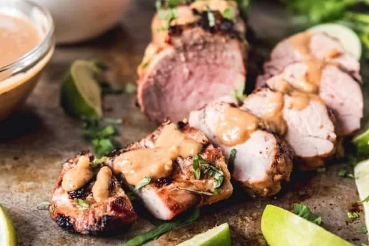 A close-up view of sliced grilled pork tenderloin topped with a creamy mustard sauce, garnished with fresh herbs. Lime wedges and a small bowl of extra sauce are placed beside the tenderloin on a rustic wooden surface.