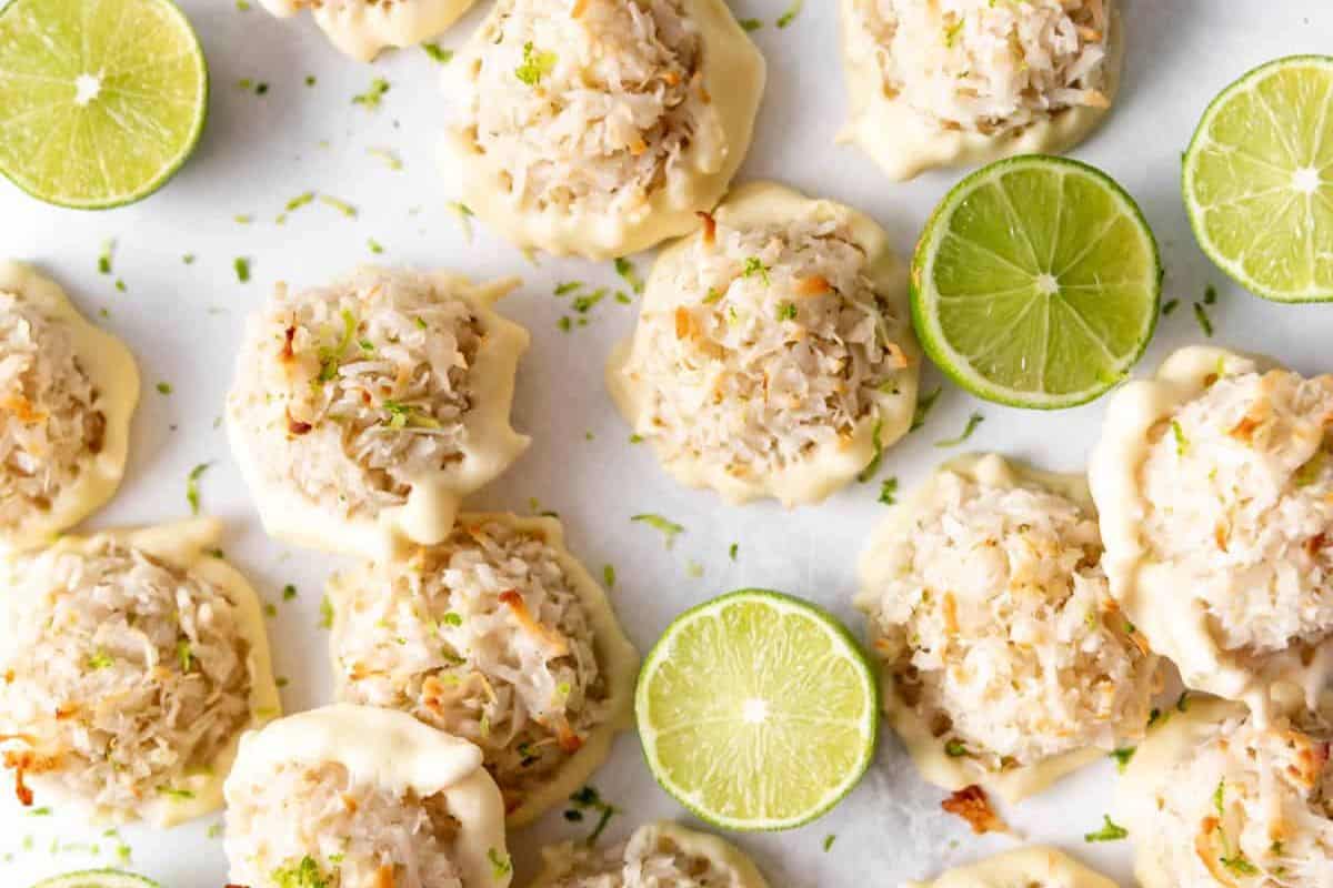 Close-up of coconut macaroons dipped in white chocolate with lime zest sprinkled on top, arranged on a white surface. Fresh lime slices are interspersed among the macaroons, adding a pop of green to the scene.