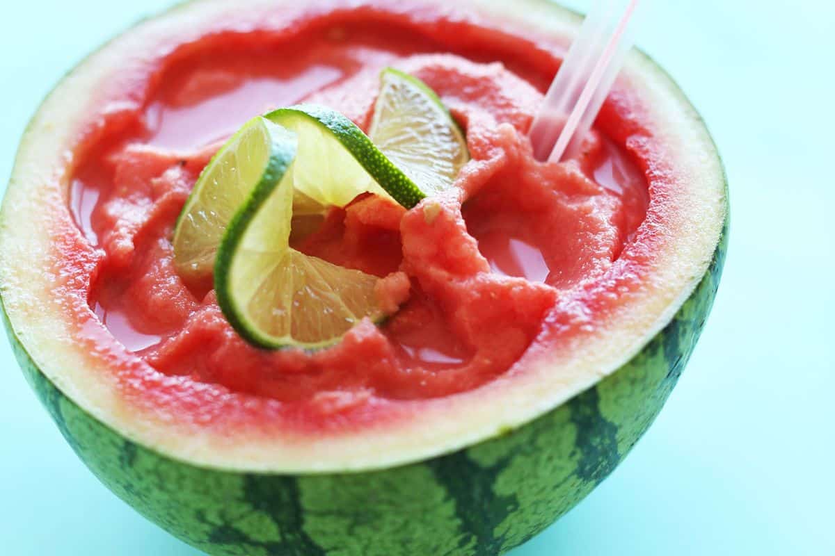A watermelon half filled with blended watermelon juice, garnished with thin slices of lime and a curly lime peel, with a straw inserted for drinking. The background is a light turquoise color.