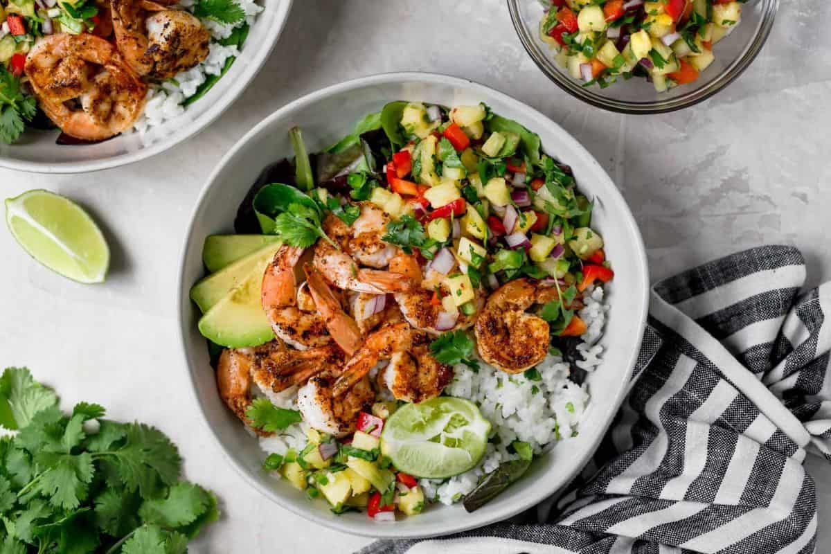A bowl filled with grilled shrimp, avocado slices, white rice, and a colorful mix of diced vegetables, including red peppers and cucumbers. A lime wedge is on top, and fresh cilantro is scattered around. A striped napkin and a smaller bowl with more diced vegetables are nearby.