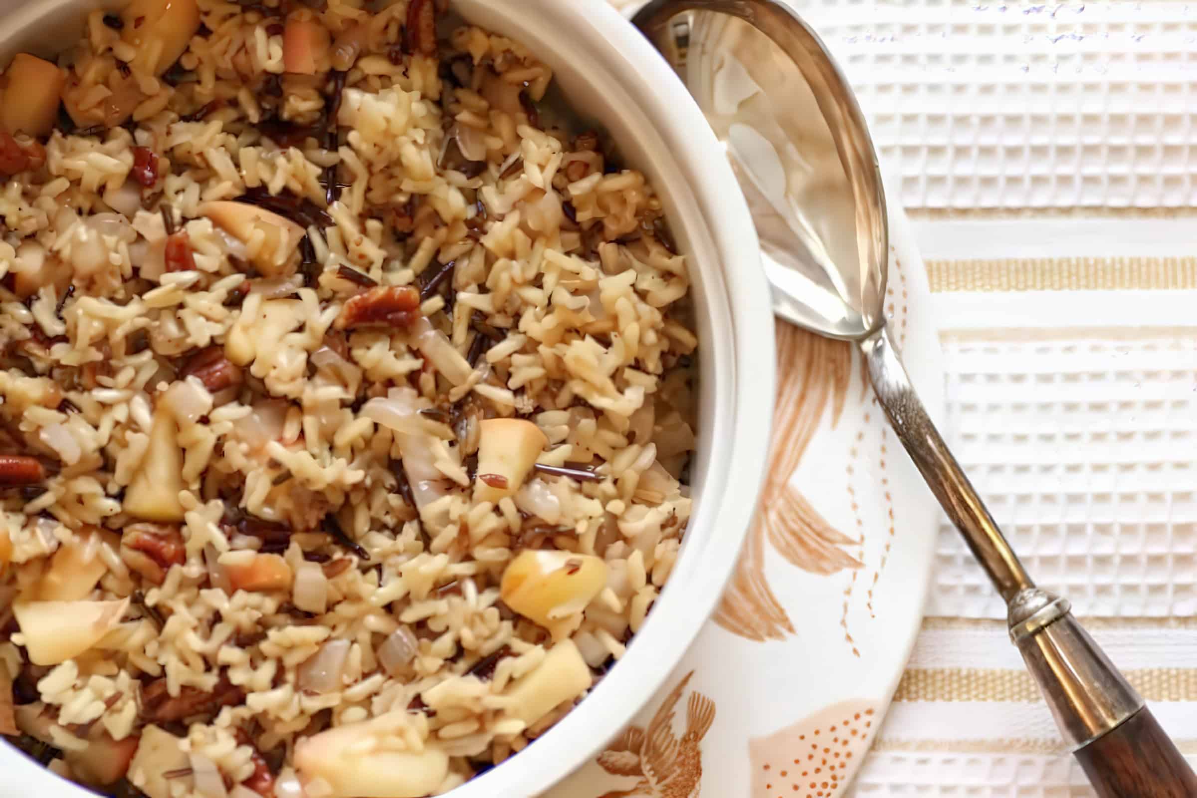A white bowl filled with a rice dish, featuring a mix of brown and wild rice, diced apples, pecans, and onions. The bowl is placed on a patterned plate with a metal spoon resting beside it on a white and gold striped napkin.