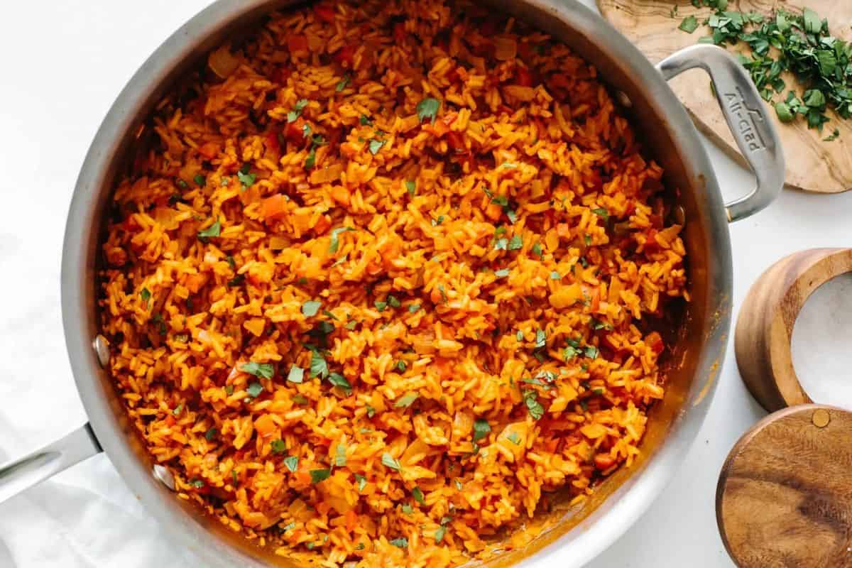 A large stainless steel pot filled with bright-orange Mexican rice garnished with chopped cilantro. The pot is surrounded by wooden containers and a cutting board with more chopped cilantro on a white surface.