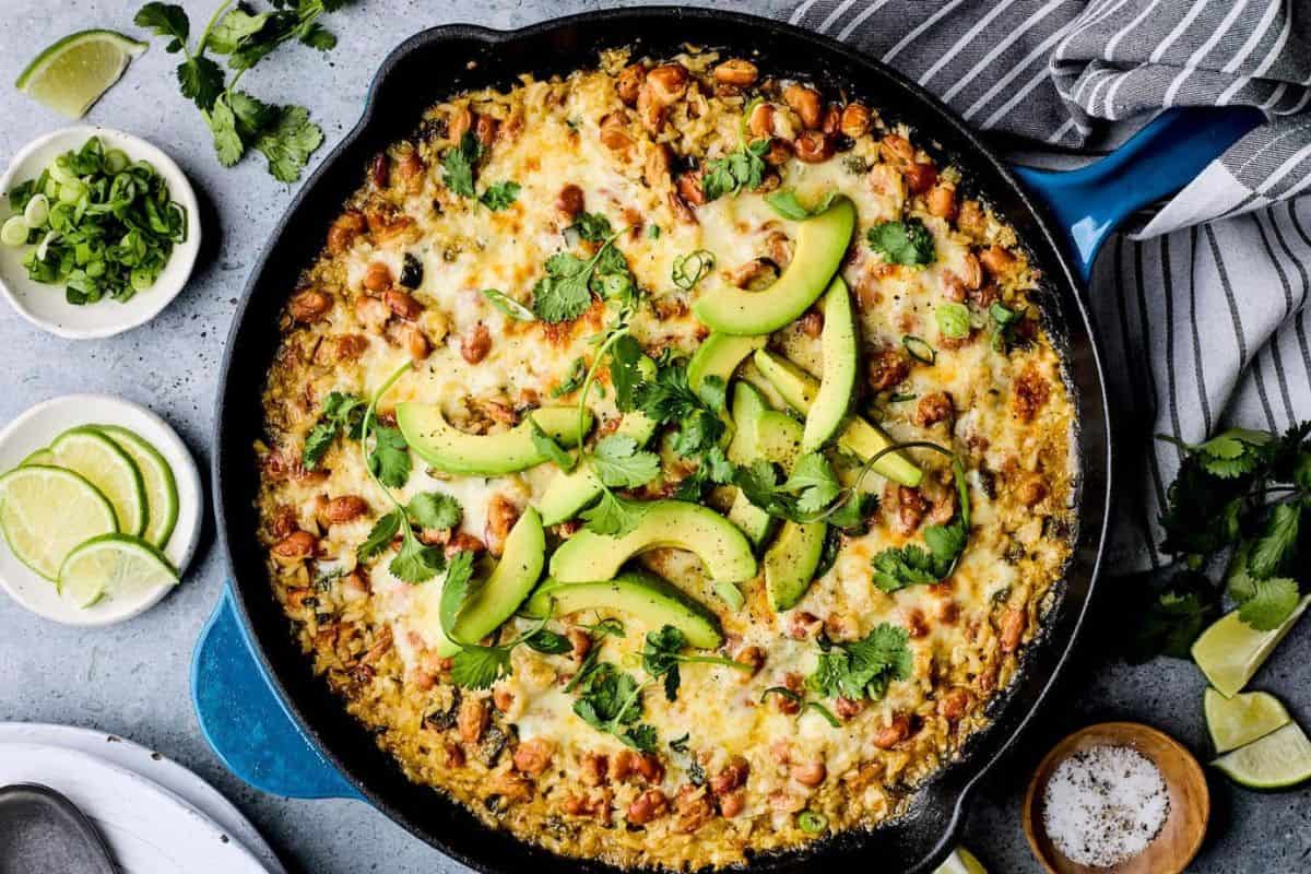 A skillet filled with cheesy chicken and rice, topped with sliced avocado and fresh cilantro. Beside the skillet are small bowls containing lime wedges, chopped green onions, and additional cilantro, along with a striped cloth and scattered cilantro leaves.