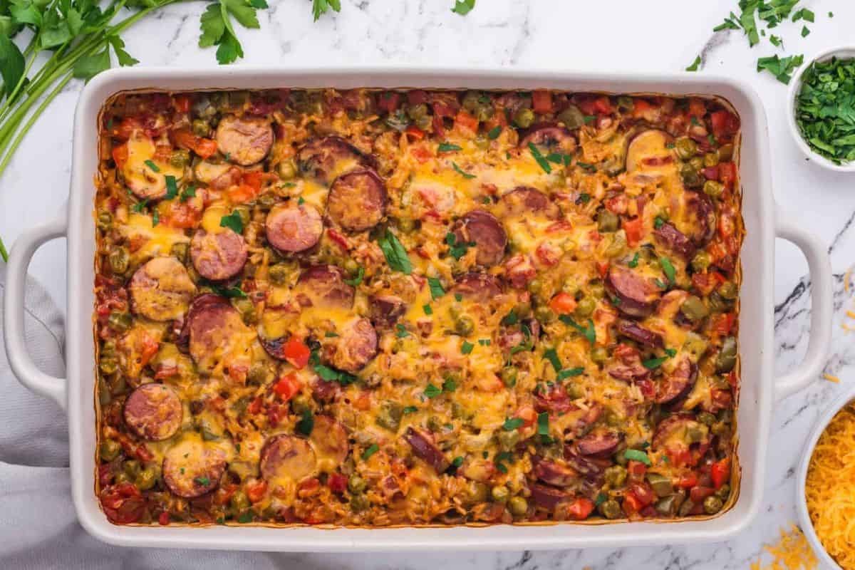 A colorful baked casserole dish filled with sliced sausages, mixed vegetables, and melted cheese, garnished with fresh parsley. The dish is set on a white marble surface with fresh parsley and a small bowl of chopped herbs nearby.
