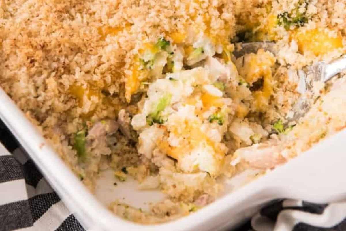A close-up of a casserole dish filled with a baked chicken and broccoli casserole. The dish contains a golden breadcrumb topping, with a serving taken out, revealing pieces of chicken, broccoli, and a creamy, cheesy mixture underneath. A serving spoon is also visible.
