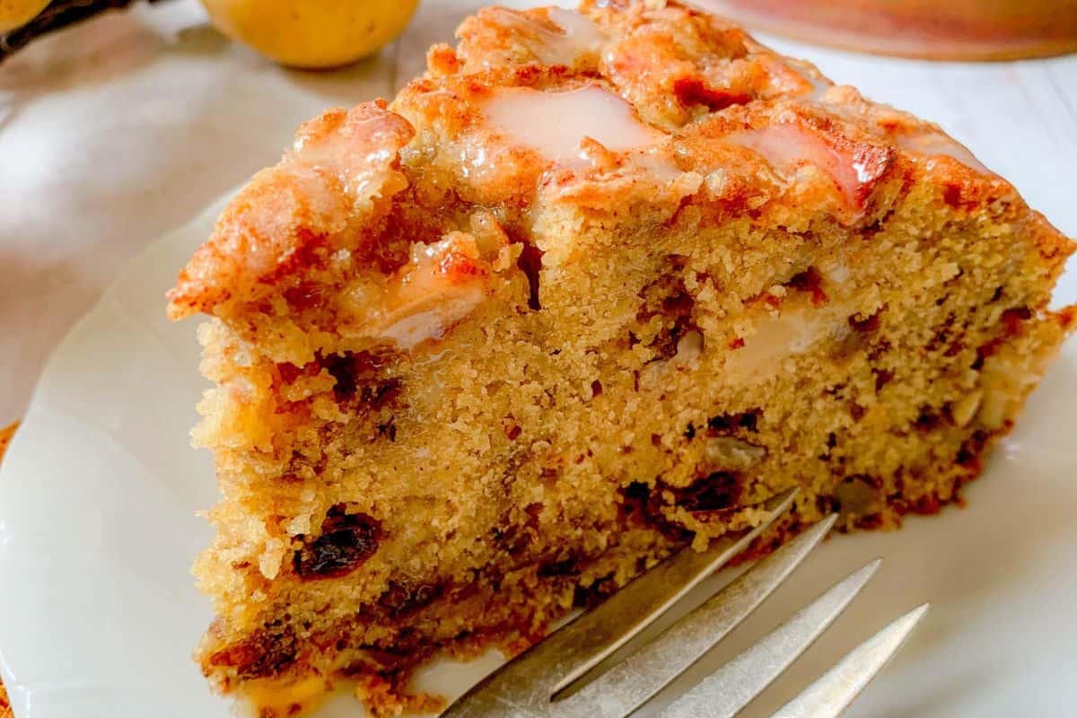 A slice of apple coffee cake with visible chunks of apple and raisins, topped with a light glaze, placed on a white plate with a fork beside it.