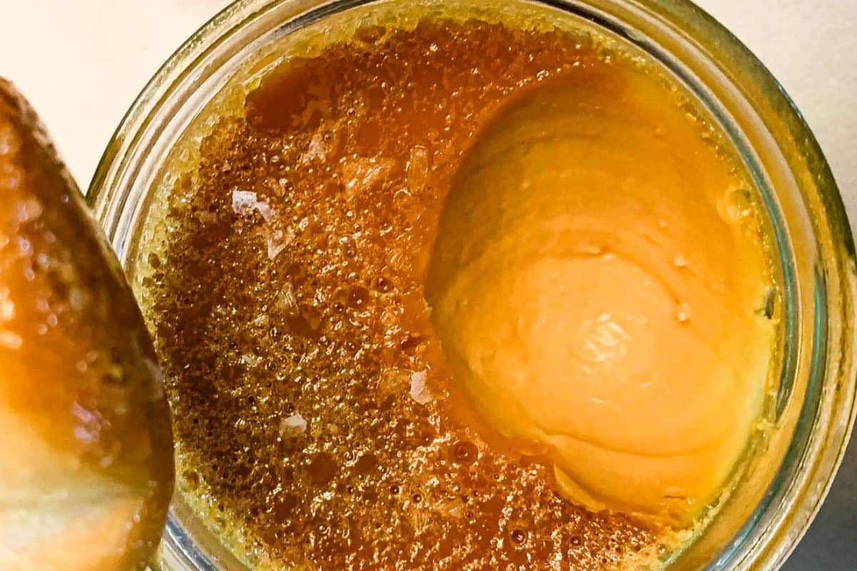 Four small glass jars filled with salted caramel custard are arranged on a wooden surface. The custard topping is glossy with a few flakes of sea salt sprinkled on top. A beige napkin with cutlery is partially visible in the background.