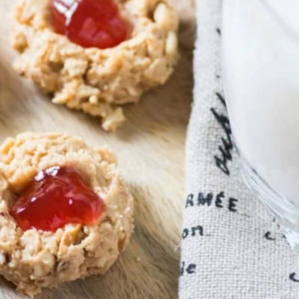 Three thumbprint cookies with red jelly centers sit next to a glass of milk on a beige cloth.