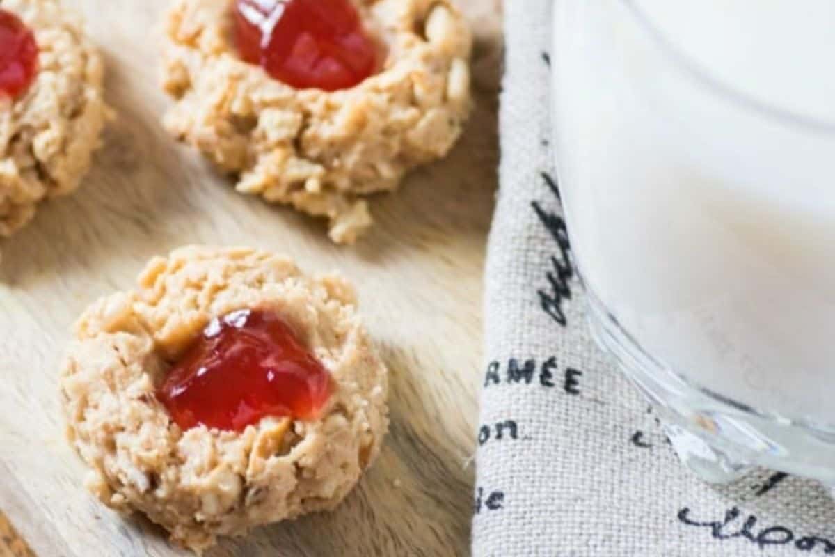 Three thumbprint cookies filled with red jam sit on a wooden surface next to a glass of milk with a cloth napkin underneath. The cookies have a rough, crumbly texture and the napkin has black printed text.