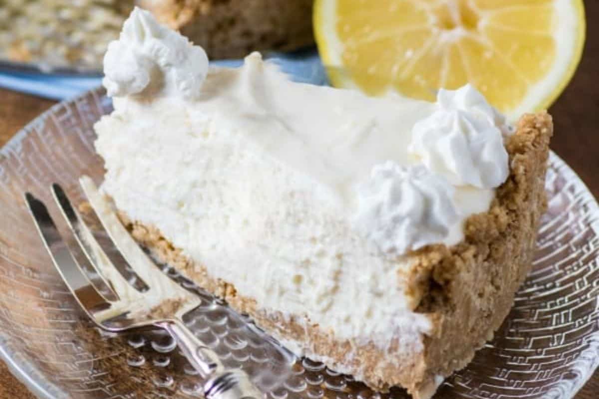 A slice of creamy cheesecake topped with dollops of whipped cream sits on a clear glass plate with a decorative fork beside it. In the background, there is a halved lemon, adding a hint of citrus to the scene.