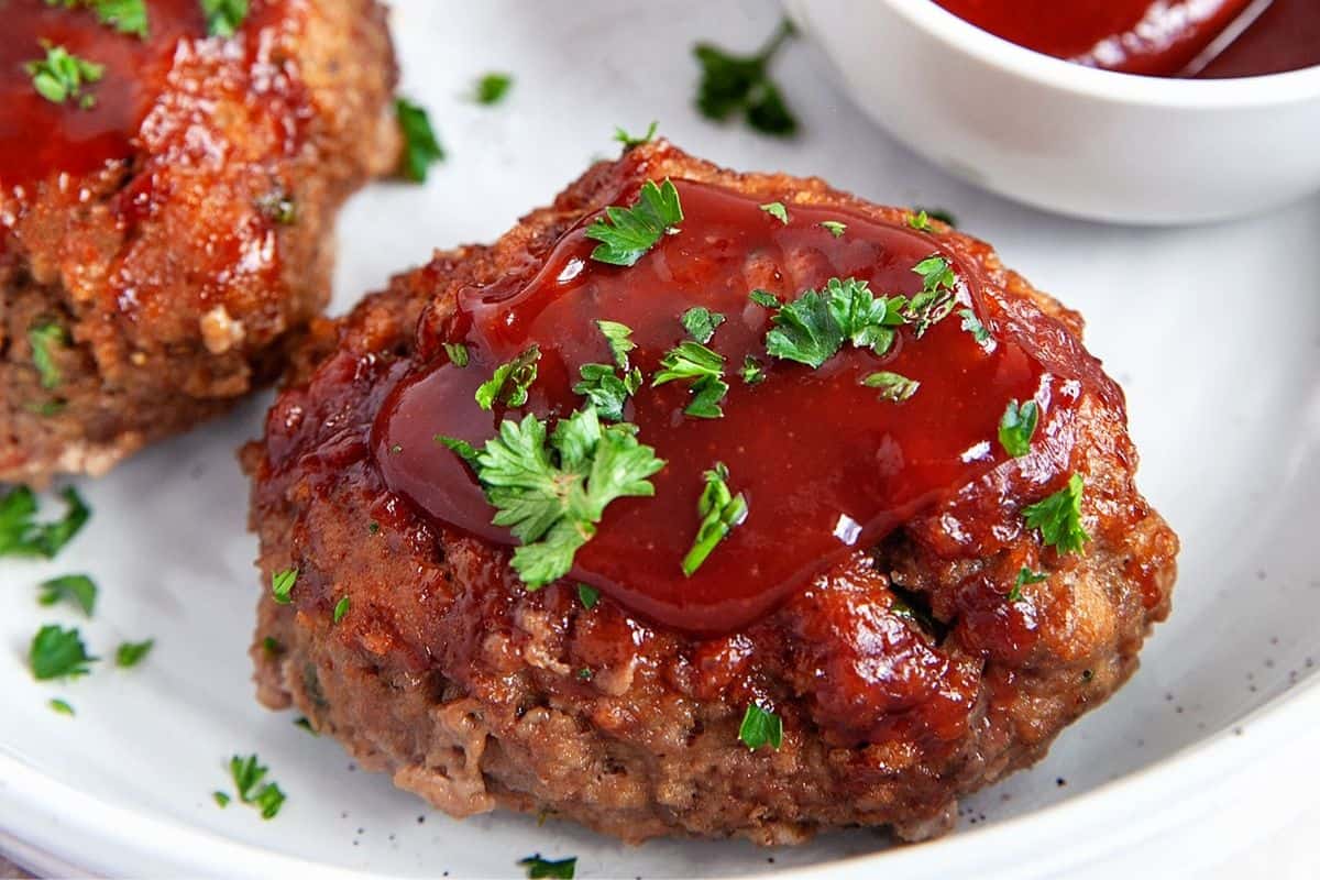 Two mini meatloaves topped with a generous drizzle of ketchup and garnished with fresh chopped parsley are placed on a white plate. A small bowl of ketchup is visible in the background.