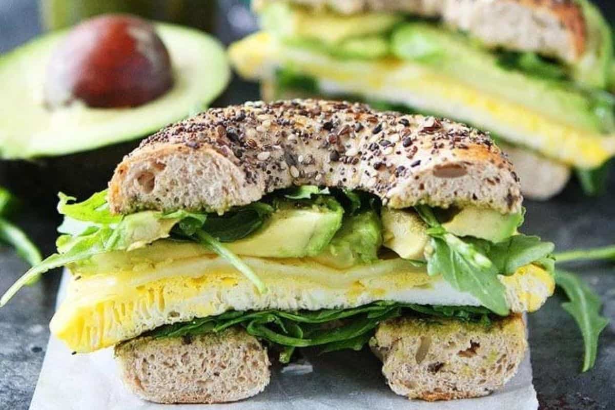 A multi-seed bagel sandwich filled with fresh spinach, slices of avocado, and a folded egg. The sandwich sits on parchment paper with a halved avocado in the background. The filling is vibrant and hearty, showcasing layers of green and yellow.A multi-seed bagel sandwich filled with fresh spinach, slices of avocado, and a folded egg. The sandwich sits on parchment paper with a halved avocado in the background. The filling is vibrant and hearty, showcasing layers of green and yellow.