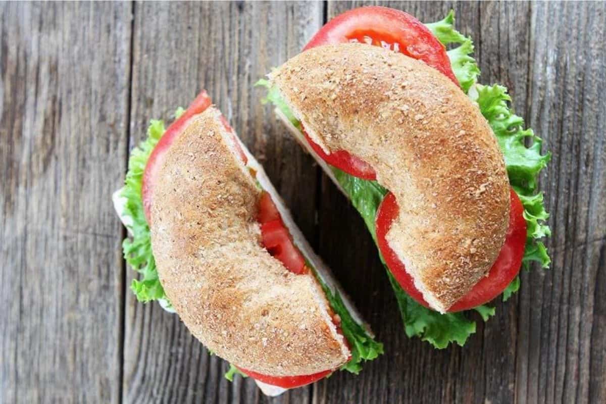 A wheat bagel sandwich cut in half and placed on a rustic wooden surface. The sandwich is filled with lettuce and tomato slices.