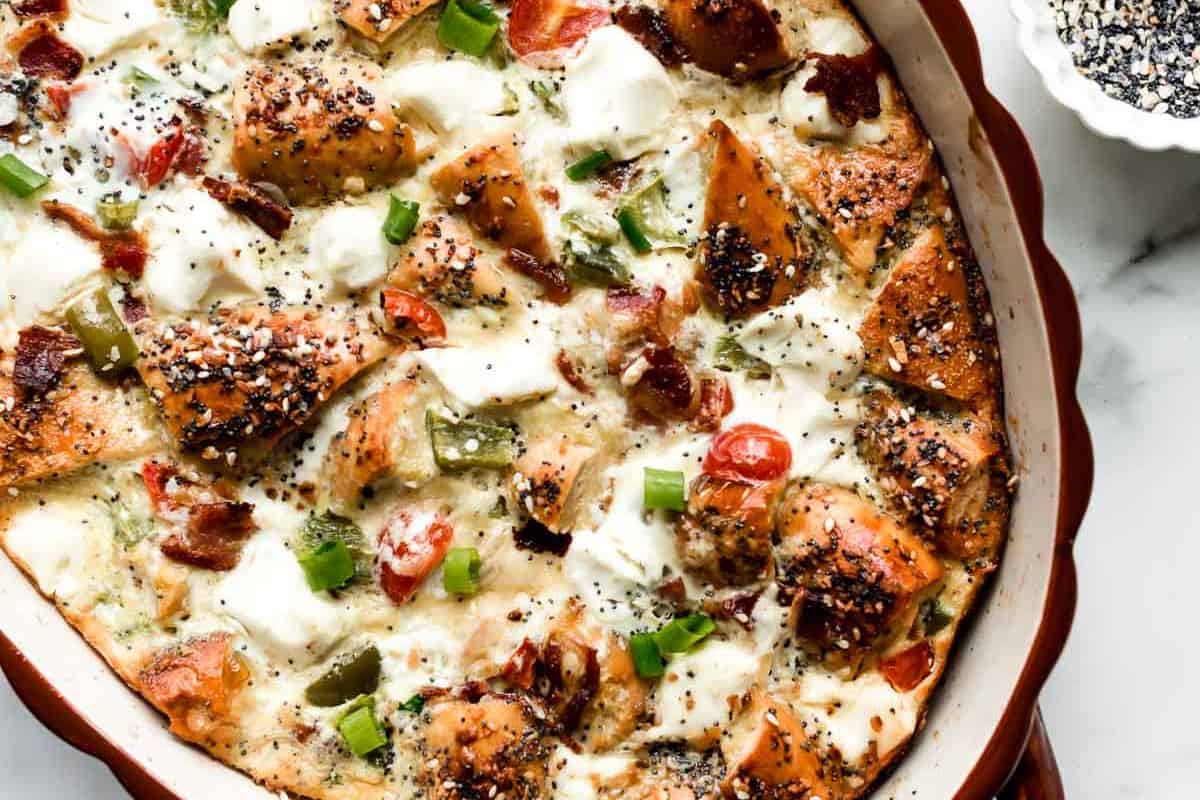 A close-up view of a savory casserole dish, featuring baked, seasoned bread cubes mixed with melted cheese, cherry tomatoes, green peppers, and bacon bits. The dish is garnished with a sprinkle of seasoning and looks crispy and golden-brown on top.