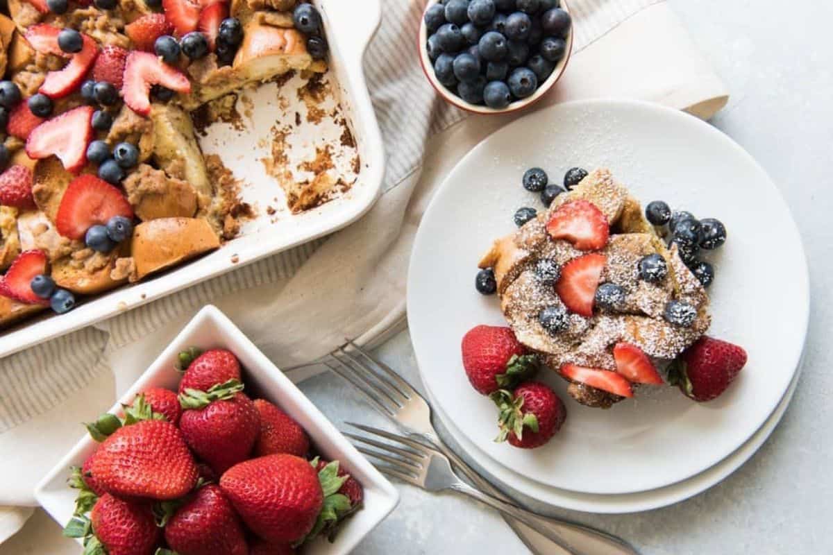A table setting features a plate of French toast casserole topped with powdered sugar, strawberries, and blueberries. Nearby, there are bowls of fresh strawberries and blueberries, a casserole dish with remaining servings, and two forks on a white cloth.