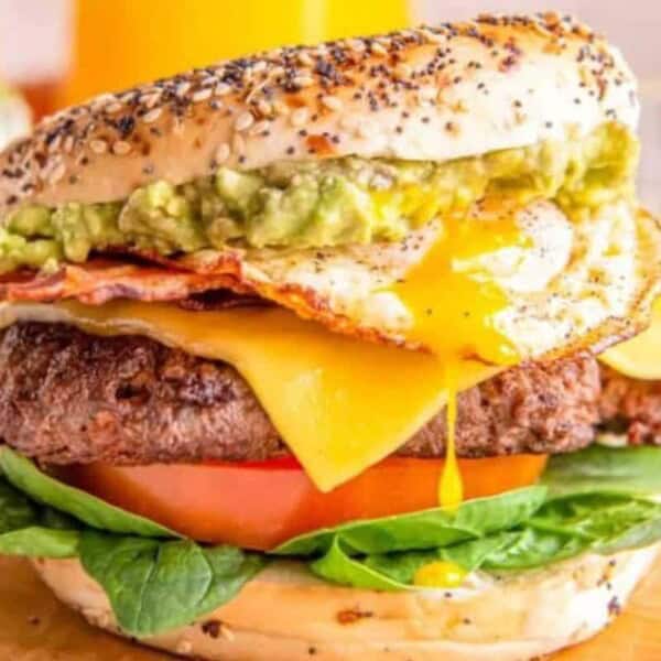 A close-up of a gourmet burger with a poppy seed bun, featuring a fried egg with a runny yolk, mashed avocado, crispy bacon, melted cheese, a beef patty, a slice of tomato, and fresh spinach leaves. The burger is placed on a wooden surface, with juice in the background.