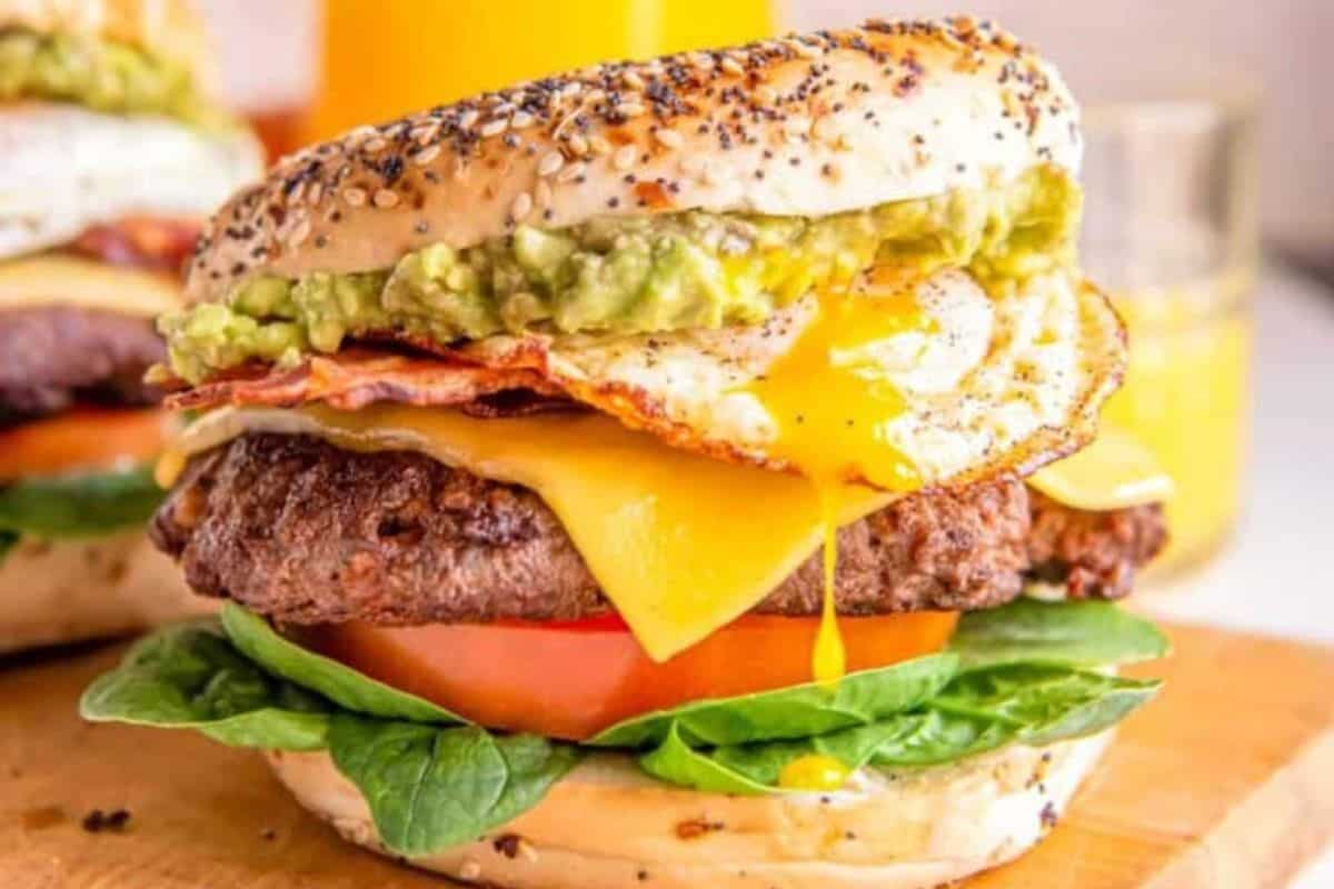 A close-up of a gourmet burger with a poppy seed bun, featuring a fried egg with a runny yolk, mashed avocado, crispy bacon, melted cheese, a beef patty, a slice of tomato, and fresh spinach leaves. The burger is placed on a wooden surface, with juice in the background.