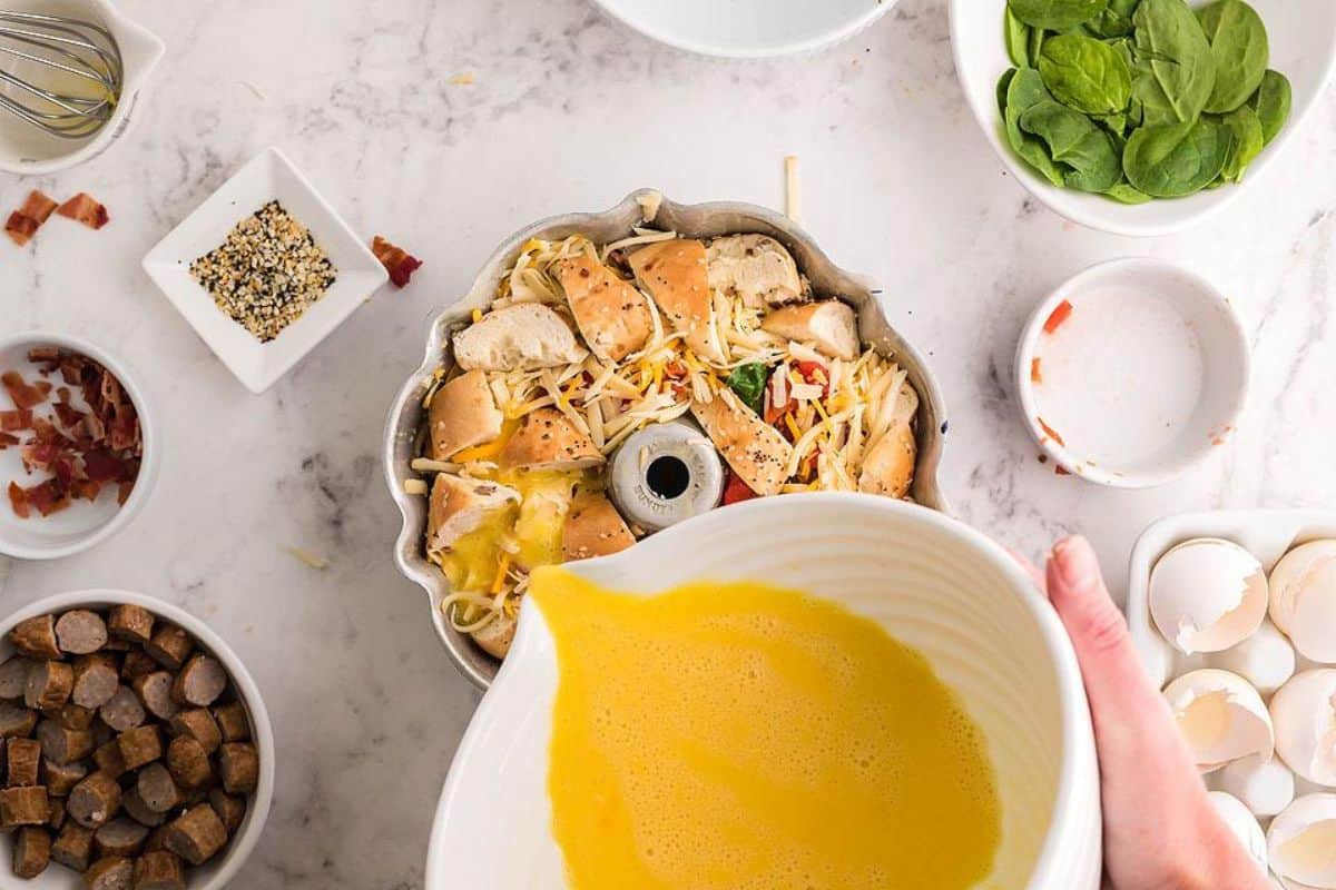 A person pours beaten eggs from a white bowl into a bundt pan filled with chunks of bread, shredded cheese, meat, and vegetables. The pan is on a white marble surface surrounded by bowls of spices, spinach, chopped meats, and cracked eggshells.