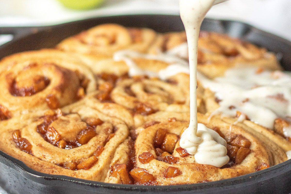 A skillet filled with freshly baked cinnamon rolls, topped with caramelized apple chunks. A thick, creamy glaze is being poured over the golden-brown rolls, partially covering them. The image captures the tempting and warm appearance of the dessert.