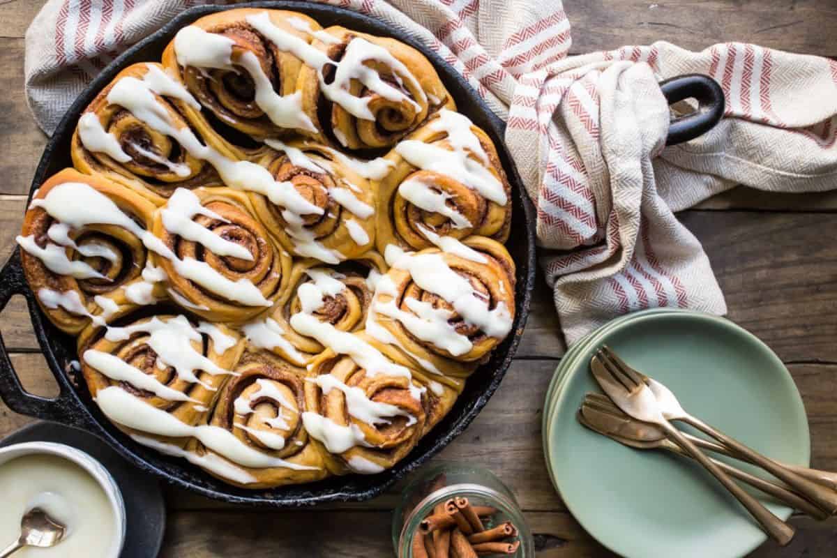 A cast iron skillet filled with freshly baked cinnamon rolls topped with vanilla icing, sitting on a wooden table. Beside the skillet, there is a striped cloth, a small jar of cinnamon sticks, an empty green plate, and gold-colored cutlery.