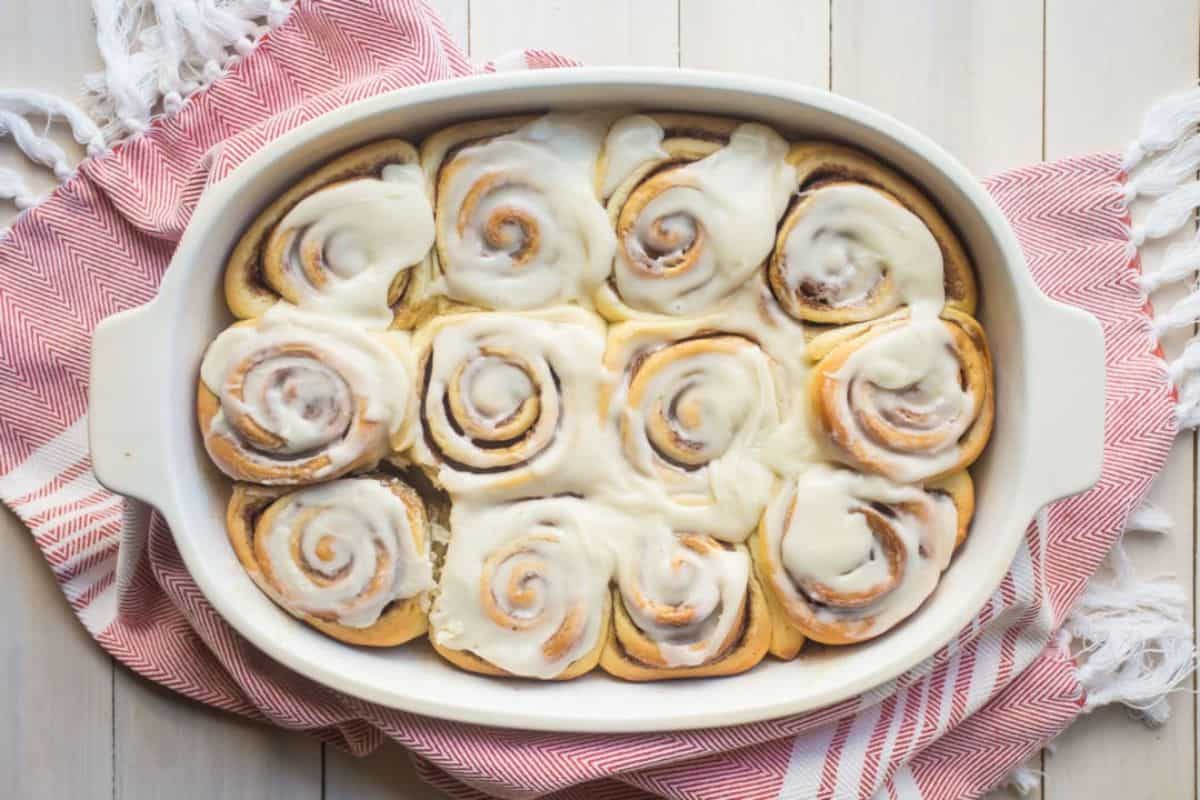 A white rectangular baking dish filled with twelve freshly baked cinnamon rolls topped with a thick layer of white icing, placed on a folded red and white striped cloth on a light wooden surface.