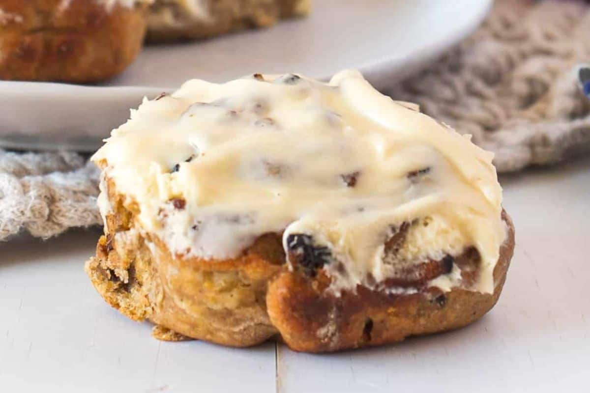 A close-up of a cinnamon roll topped with creamy white icing, resting on a light-colored surface. The roll has visible swirls of cinnamon and sugary filling, and hints of raisins or nuts can be seen peeking through the dough.
