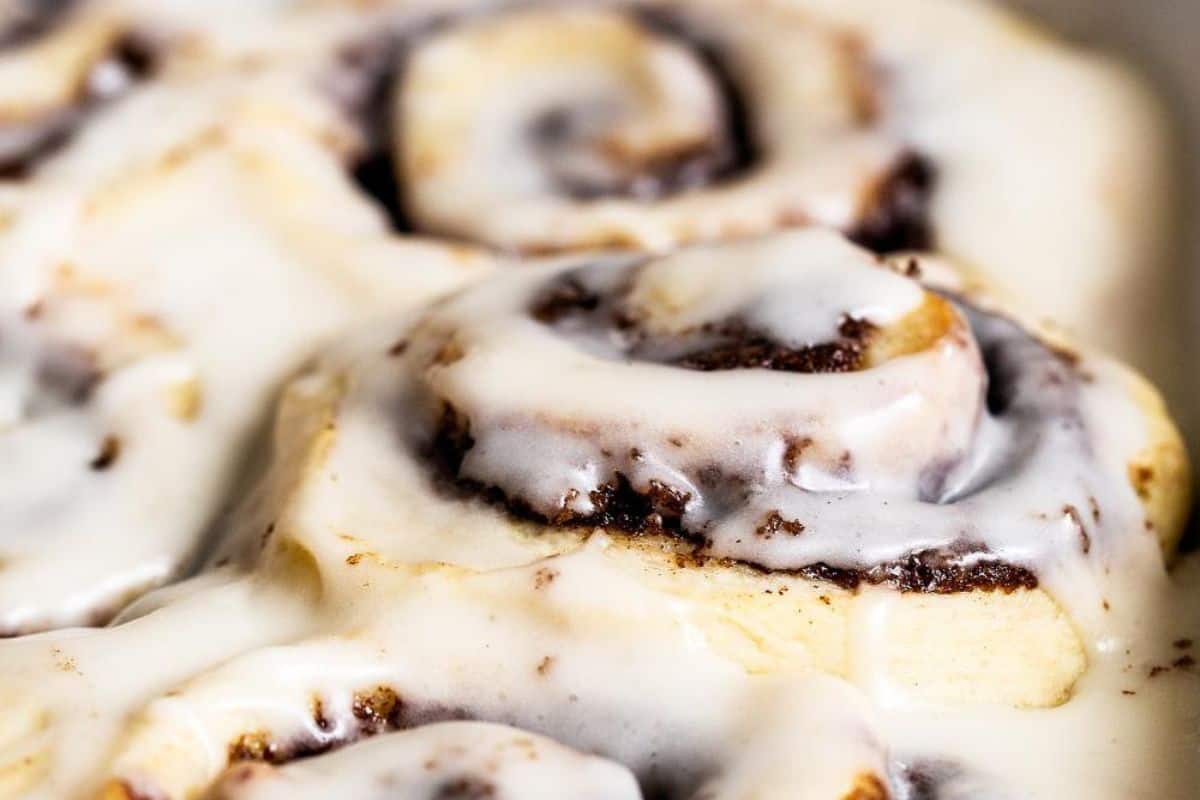 Close-up of freshly baked cinnamon rolls covered in a creamy white icing. The rolls appear soft and fluffy with a visible swirl of brown cinnamon filling peeking through the glaze. The icing is applied generously, dripping slightly over the sides of the rolls.