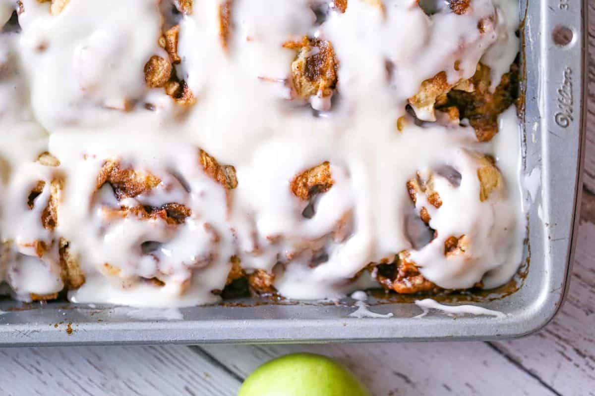 A close-up of a baking tray filled with freshly baked cinnamon rolls covered in white icing. One green apple is partially visible in the bottom right corner of the image, placed on a wooden surface.