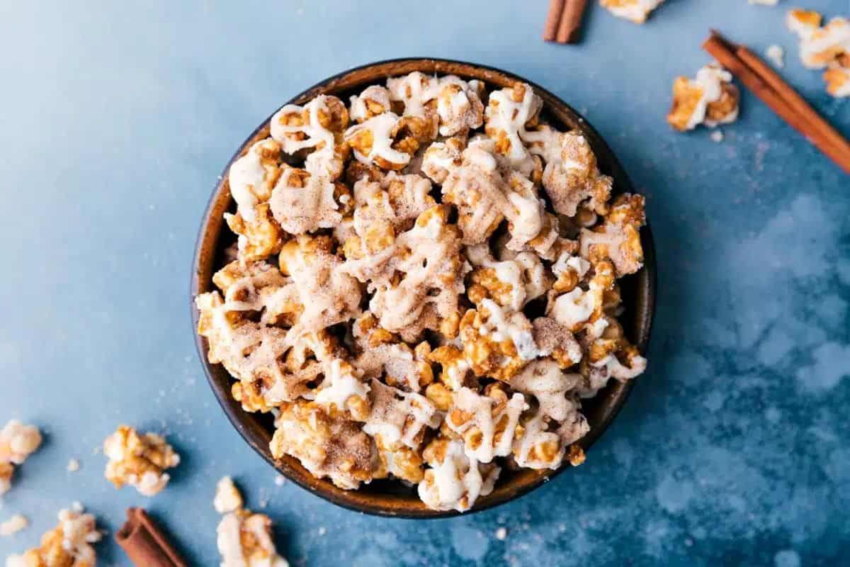 A bowl of caramel popcorn is drizzled with white chocolate and sprinkled with cinnamon on a blue background. Loose popcorn pieces and cinnamon sticks are scattered around the bowl.