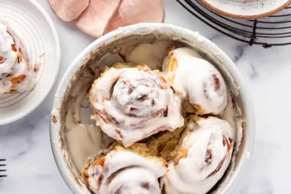A bowl filled with six frosted cinnamon rolls sits on a marble surface. A small plate with a single cinnamon roll is placed nearby, alongside a wire rack and a pink cloth in the background. The cinnamon rolls are generously coated with icing.