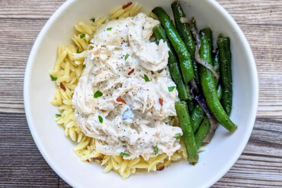 A close-up of shredded chicken in a creamy lemon sauce, garnished with lemon slices and sprinkled with herbs. The dish is presented in a black bowl, showcasing a rich and savory appearance.
