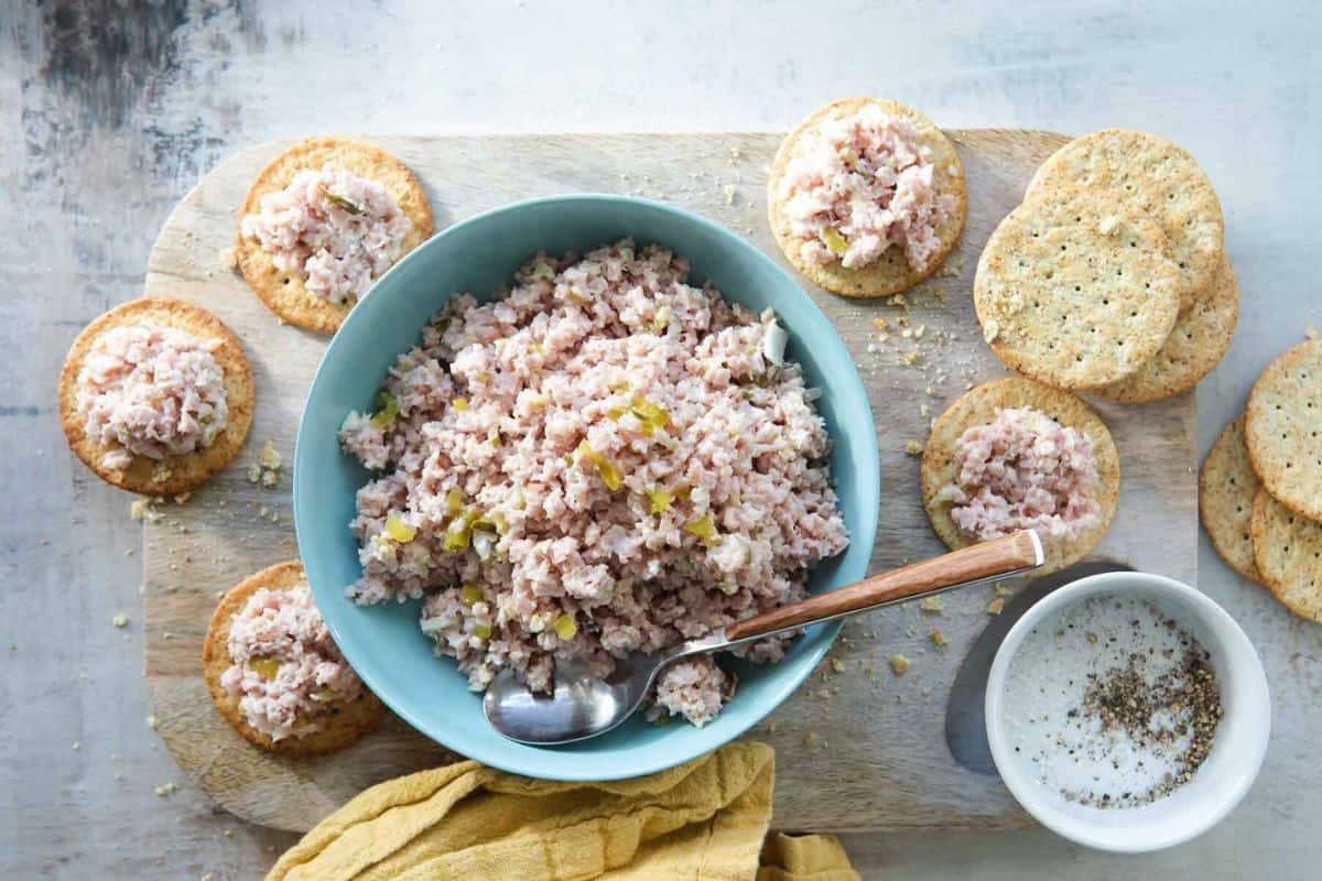 A bowl of ham salad with a wooden spoon sits on a cutting board, surrounded by round crackers topped with ham salad. A small bowl of pepper-dusted mayo is on the side, along with a yellow cloth. Additional plain crackers are scattered around.