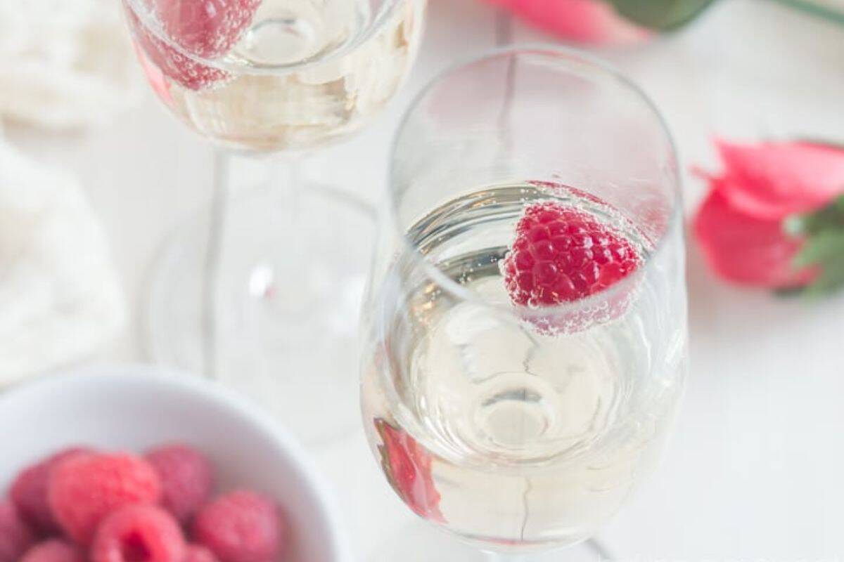 Two glasses of clear sparkling wine, each garnished with a raspberry, are on a light-colored table. In the foreground, there is a white bowl filled with additional raspberries. Pink roses are faintly visible in the background.